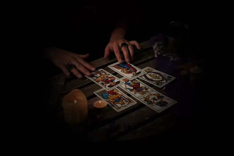 Il y a six cartes en quinconce sur une table. La photo est sombre est elle montre les mains d'une voyante pendent le tirage. Sur une table en bois les cartes sont étalés et on voit deux bougies à gauche.