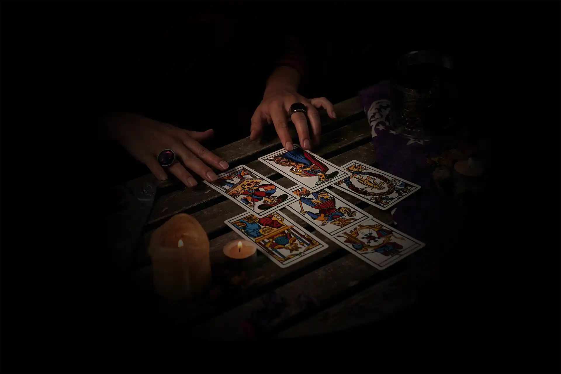 Il y a six cartes en quinconce sur une table. La photo est sombre est elle montre les mains d'une voyante pendent le tirage. Sur une table en bois les cartes sont étalés et on voit deux bougies à gauche.